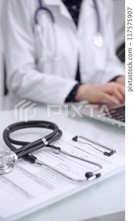 Stethoscope is lying on the glass table while doctor woman is working with laptop computer on the background, close-up. Medicine and pharmacy concept 117575907