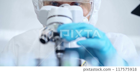 Professional scientist in protective gear is using microscope in a laboratory setting, close-up of researcher. Science and medicine concepts Professional scientist in protective gear is using microscope in a laboratory setting, close-up of researcher. Science and medicine concepts 117575918