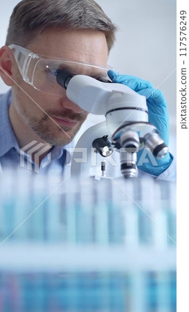 Man scientist with protective glasses and gloves is using microscope in laboratory, vertical portrait of researcher. Microbiology science concept Man scientist with protective glasses and gloves is using microscope in laboratory, vertical portrait of researcher. Microbiology science concept 117576249