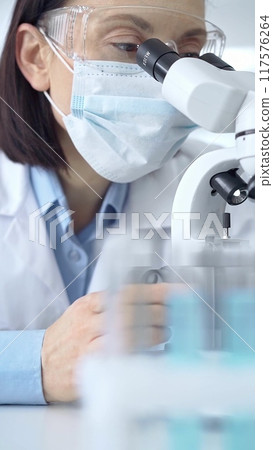 Female scientist using microscope in laboratory. Focus on a woman in lab goggles analyzing samples with a microscope in a scientific setting 117576264