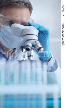 Man scientist with protective glasses, mask and gloves is using microscope in laboratory, vertical portrait of researcher. Microbiology and science Man scientist with protective glasses, mask and gloves is using microscope in laboratory, vertical portrait of researcher. Microbiology and science 117576265
