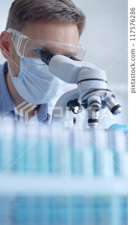 Man scientist with protective glasses, mask and gloves is using microscope in laboratory, vertical portrait of researcher. Microbiology and science Man scientist with protective glasses, mask and gloves is using microscope in laboratory, vertical portrait of researcher. Microbiology and science 117576286