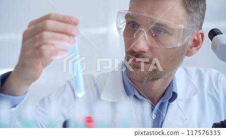 Scientist researcher with protective glasses examining lab tube with a blue fluid. Microbiology and science concept 117576335