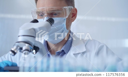 Man scientist with protective glasses, mask and gloves is using microscope in laboratory, portrait of researcher. Microbiology science concept Man scientist with protective glasses, mask and gloves is using microscope in laboratory, portrait of researcher. Microbiology science concept 117576356