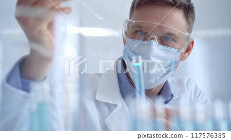 Man with safety glasses and blue mask is working with lab tubes using a pipette in a modern laboratory, close-up of researcher. Science and medicine Man with safety glasses and blue mask is working with lab tubes using a pipette in a modern laboratory, close-up of researcher. Science and medicine 117576385