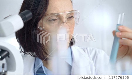 Female scientist analyzing sample in laboratory. Focused female researcher examines a clear test tube in a modern lab setting Female scientist analyzing sample in laboratory. Focused female researcher examines a clear test tube in a modern lab setting 117576396