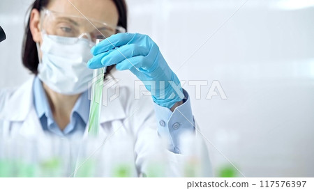 Woman scientist with protective gloves and glasses examining lab tube with a green fluid. Microbiology and science concept Woman scientist with protective gloves and glasses examining lab tube with a green fluid. Microbiology and science concept 117576397