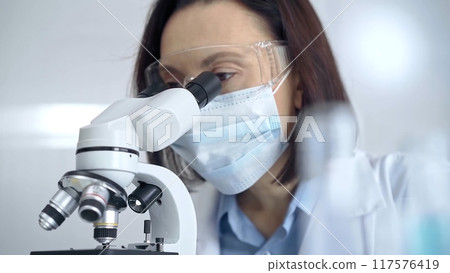 Female scientist in lab goggles and protective mask using microscope in laboratory Female scientist in lab goggles and protective mask using microscope in laboratory 117576419