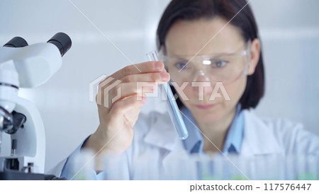 Scientist female researcher with protective glasses examining lab tube with a blue fluid. Microbiology and science concept 117576447