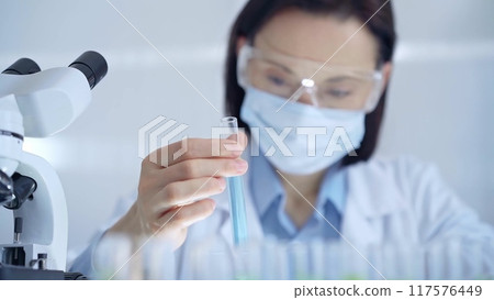 Scientist female researcher with protective glasses and mask examining lab tube with a blue fluid. Microbiology and science concept Scientist female researcher with protective glasses and mask examining lab tube with a blue fluid. Microbiology and science concept 117576449