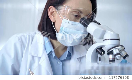 Female scientist in lab goggles, protective mask and gloves is using microscope in laboratory. Science concept 117576508