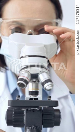 Scientist using microscope in laboratory. Close up on a woman face with a lab goggles in a scientific setting 117576514