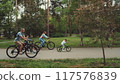 Family enjoying bike ride in park, with parents and children cycling together on paved path surrounded by trees. Concept of family bonding, outdoor activities, and healthy lifestyle 117576839