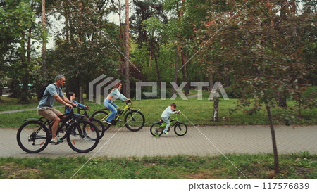 Family enjoying bike ride in park, with parents and children cycling together on paved path surrounded by trees. Concept of family bonding, outdoor activities, and healthy lifestyle Family enjoying bike ride in park, with parents and children cycling together on paved path surrounded by trees. Concept of family bonding, outdoor activities, and healthy lifestyle 117576839