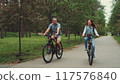 Couple enjoys biking together in park, smiling and talking while riding on tree-lined path. Concept of outdoor activities, bonding, and healthy lifestyle 117576840