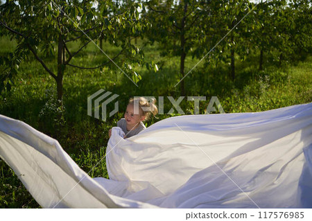a family of four is playing with a white sheet in the garden, a toddler wants to flutter the sheet 117576985