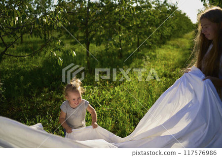 a family of four is playing with a white sheet in the garden, a toddler wants to flutter the sheet 117576986