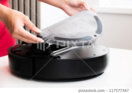 Smart round vacuum cleaner in female hands. Woman takes out container and filter to clean it from dirt and debris. Concept of cleaning with cordless vacuum cleaner, close-up 117577367