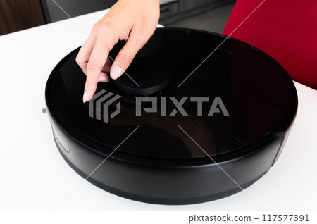 Robot vacuum cleaner with round black body on white background table, new home assistant, woman hand reaches out to turn it on and check, close-up Robot vacuum cleaner with round black body on white background table, new home assistant, woman hand reaches out to turn it on and check, close-up 117577391