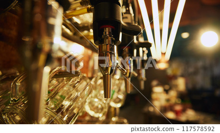 Close-up of multiple shiny beer taps with handles and mirrors, set against blurred background with rows of empty glasses and bar counter in the background. Close-up of multiple shiny beer taps with handles and mirrors, set against blurred background with rows of empty glasses and bar counter in the background. 117578592