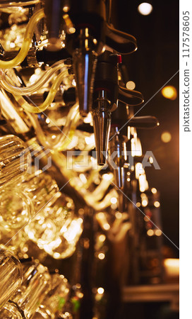 Close-up of multiple shiny beer taps with handles, set against blurred background with rows of empty glasses hanging from it. Cozy lights 117578605