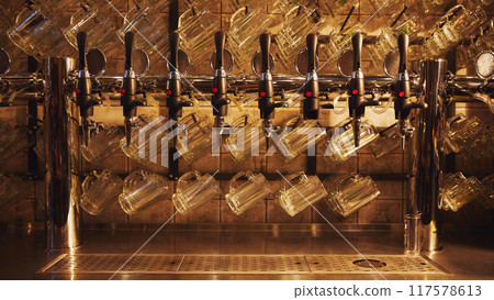 Row of shiny beer taps with handles, set against wall with rows of empty mugs hanging from it. 117578613