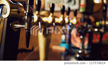 Close-up of row of shiny beer taps with handles, set against dark background with blurred bar counter. Vintage restaurant aesthetics 117578624