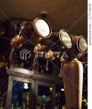 Close-up of row of shiny brass beer taps with handles and mirrors, set against dark background with blurred bar counter. Vintage bar aesthetic 117578630