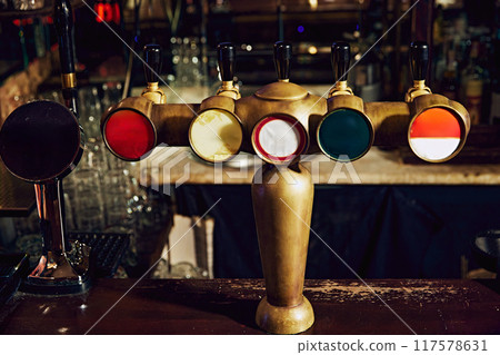 Close-up of row of beer taps with colorful handles and mirrors, set against dark background with blurred bar counter and empty glasses in background. 117578631