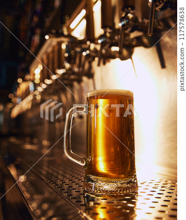 Close-up of frosty mug of golden beer with bubbles, sitting on metallic counter in front of row of beer taps. 117578638