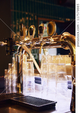 Close-up of shiny beer tap with multiple handles, set against dark background with empty glasses and metal grid in the foreground. 117578665
