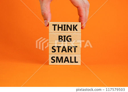 Think big start small symbol. Concept words 'Think big start small' on wooden blocks on a beautiful orange background. Businessman hand. Business, motivational and think big start small concept. Think big start small symbol. Concept words 'Think big start small' on wooden blocks on a beautiful orange background. Businessman hand. Business, motivational and think big start small concept. 117579503