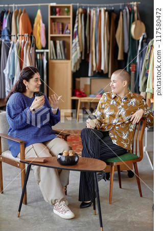 Vertical shot of two young female friends dressed in vintage outfits chatting over cup of coffee cheerfully discussing fashion while sitting in second hand chairs at thrift store, copy space Vertical shot of two young female friends dressed in vintage outfits chatting over cup of coffee cheerfully discussing fashion while sitting in second hand chairs at thrift store, copy space 117580472