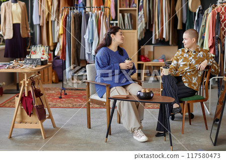 Wide angle shot of two Gen Z female friends sitting at tea table in vintage chairs chatting over cup of coffee discussing fashion at thrift store, copy space Wide angle shot of two Gen Z female friends sitting at tea table in vintage chairs chatting over cup of coffee discussing fashion at thrift store, copy space 117580473