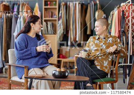 Side view of two young female friends dressed in vintage outfits chatting over cup of coffee discussing fashion while sitting in second hand chairs at thrift store, copy space 117580474