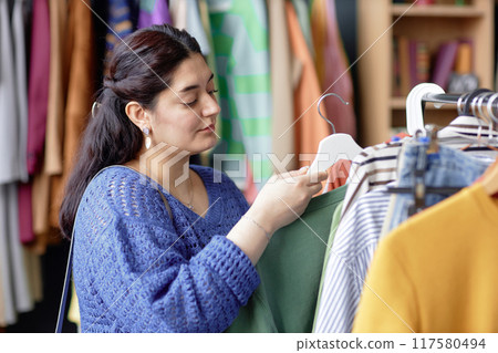 Portrait shot of young Middle Eastern woman in knitted sweater holding hanger with garment while shopping at clothes store or boutique, copy space 117580494