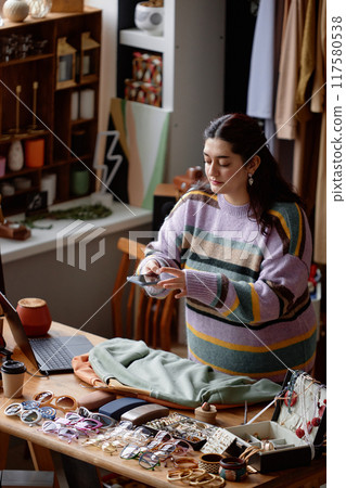 Vertical shot of Middle Eastern female sales assistant at display table taking photo with phone of second hand sweaters promoting clothes on thrift store social networks, copy space 117580538