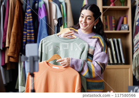 Medium shot of smiling female sales assistant of Middle Eastern ethnicity recording video on phone for account in social networks showing second hand sweaters at thrift store, copy space 117580566