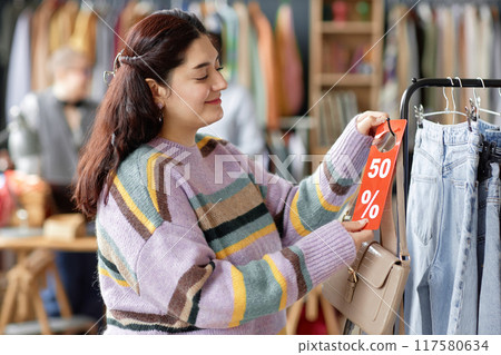 Side view medium shot of smiling Middle Eastern female customer choosing to buy second hand purse with fifty percent discount tag at thrift store Side view medium shot of smiling Middle Eastern female customer choosing to buy second hand purse with fifty percent discount tag at thrift store 117580634