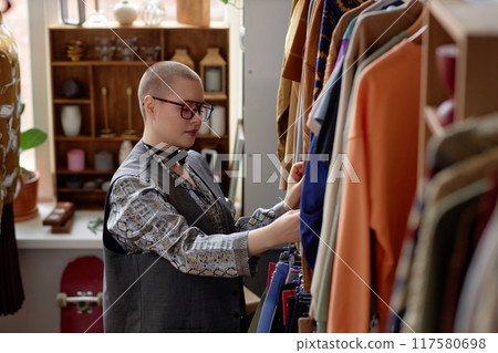 Side view of young female customer with buzz cut in vintage clothes thrifting at second hand shop choosing garments, copy space 117580698
