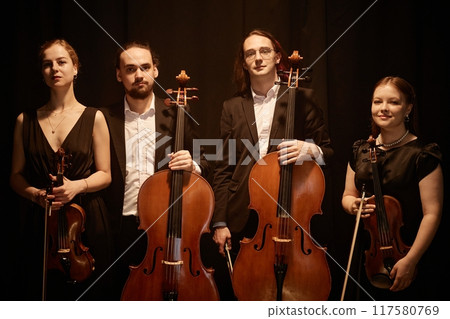 Group portrait of classical of string quartet looking at camera while standing on stage with warm top light against black curtains 117580769