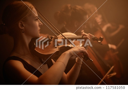 Side portrait of virtuoso female classic music artist playing violin with string quartet on stage in misty orange light 117581050