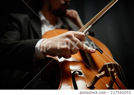 Close up on hand of unrecognizable male classical music artist strumming cello in contrast light on black background, focus on fingers and strings 117581055