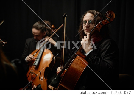 Concentrated male cello player in glasses performing classical music with string quartet in studio with dramatic top light against black background Concentrated male cello player in glasses performing classical music with string quartet in studio with dramatic top light against black background 117581057