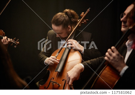 Medium shot of male musician looking down, while strumming cello playing classical music with dedication in string ensemble in studio with contrast top light on black background Medium shot of male musician looking down, while strumming cello playing classical music with dedication in string ensemble in studio with contrast top light on black background 117581062