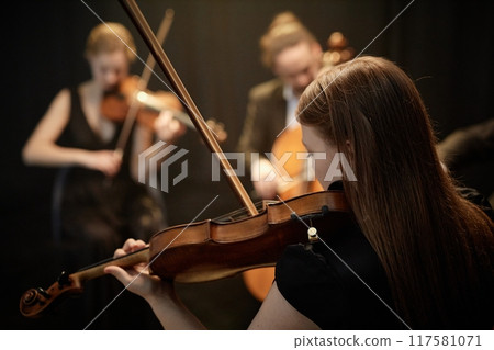 Rear view of unrecognizable female orchestra musician playing violin with string quartet in spotlight on stage with black curtains 117581071