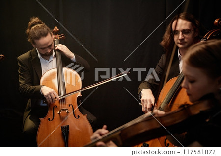 Three symphonic orchestra musicians playing cellos and violins while sitting in circle performing classical music against black curtains in contrast light Three symphonic orchestra musicians playing cellos and violins while sitting in circle performing classical music against black curtains in contrast light 117581072