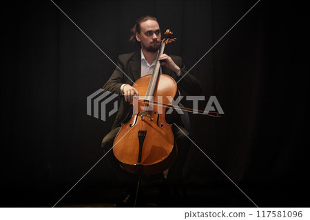 Full shot of male classical music performer playing cello during solo concert while sitting in chair against black curtains under projector light, copy space Full shot of male classical music performer playing cello during solo concert while sitting in chair against black curtains under projector light, copy space 117581096