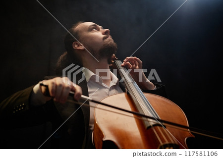 Low angle view of moved male musician thoughtfully looking up while playing cello in hazy dark studio with top light, copy space Low angle view of moved male musician thoughtfully looking up while playing cello in hazy dark studio with top light, copy space 117581106