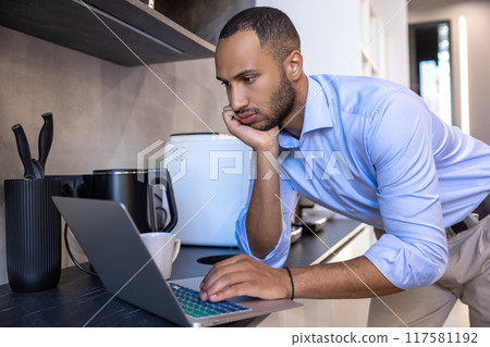 African American man wearing blue shirt working on laptop in kitchen 117581192